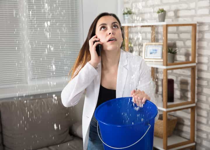 A woman calling water damage repair experts while holding a bucket trying to catch the water leaking from her ceiling in Queens, NY